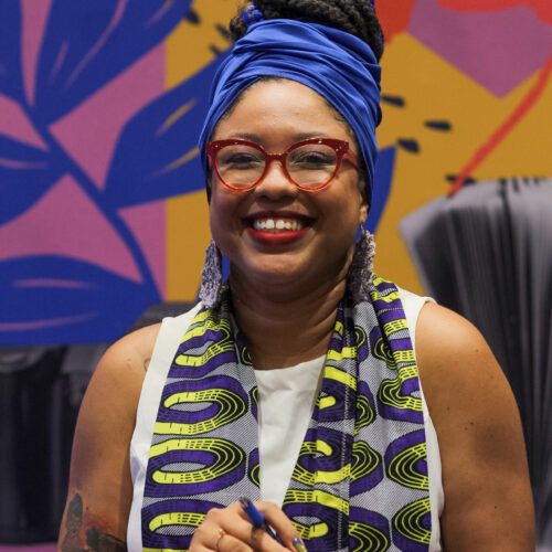 Foto da autora Lorena Pinheiro. Uma mulher negra de pele clara, com cabelo crespo preso em coque no alto da cabeça, com um turbante azul. Ela sorri para a foto, usa óculos e batom vermelho. Está com uma bata branca e um lenço com grafismos em azul e amarelo.