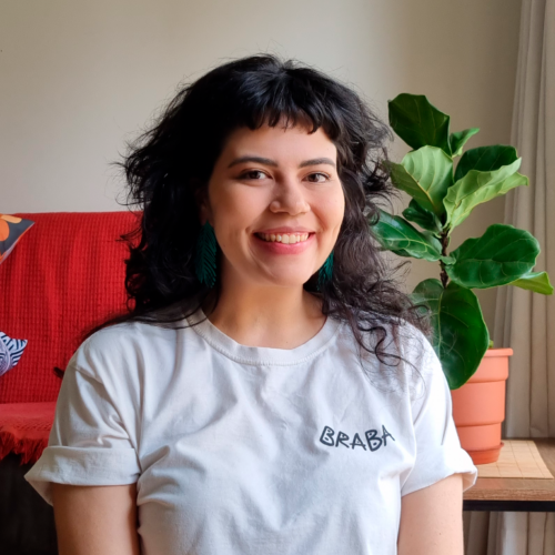 Foto de autora de Karipola. Ela é uma mulher parda, está sorrindo para a foto. Tem cabelo preto com franja no meio da testa, usa uma blusa branca. Ao fundo está um sofá vermelho e um vaso de plantas.